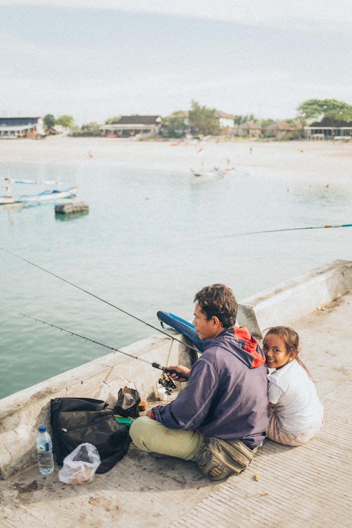 A father and daughter enjoy a peaceful fishing moment by the seaside on a sunny day.
