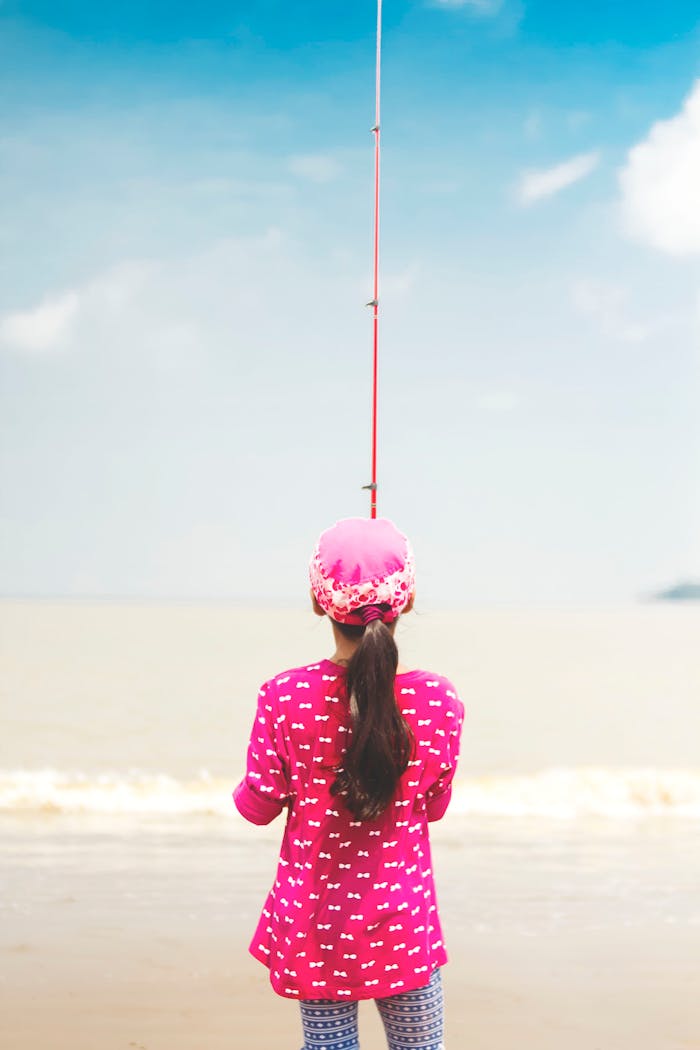 Young girl fishing at the seashore in summer, wearing pink attire, enjoying outdoor leisure.